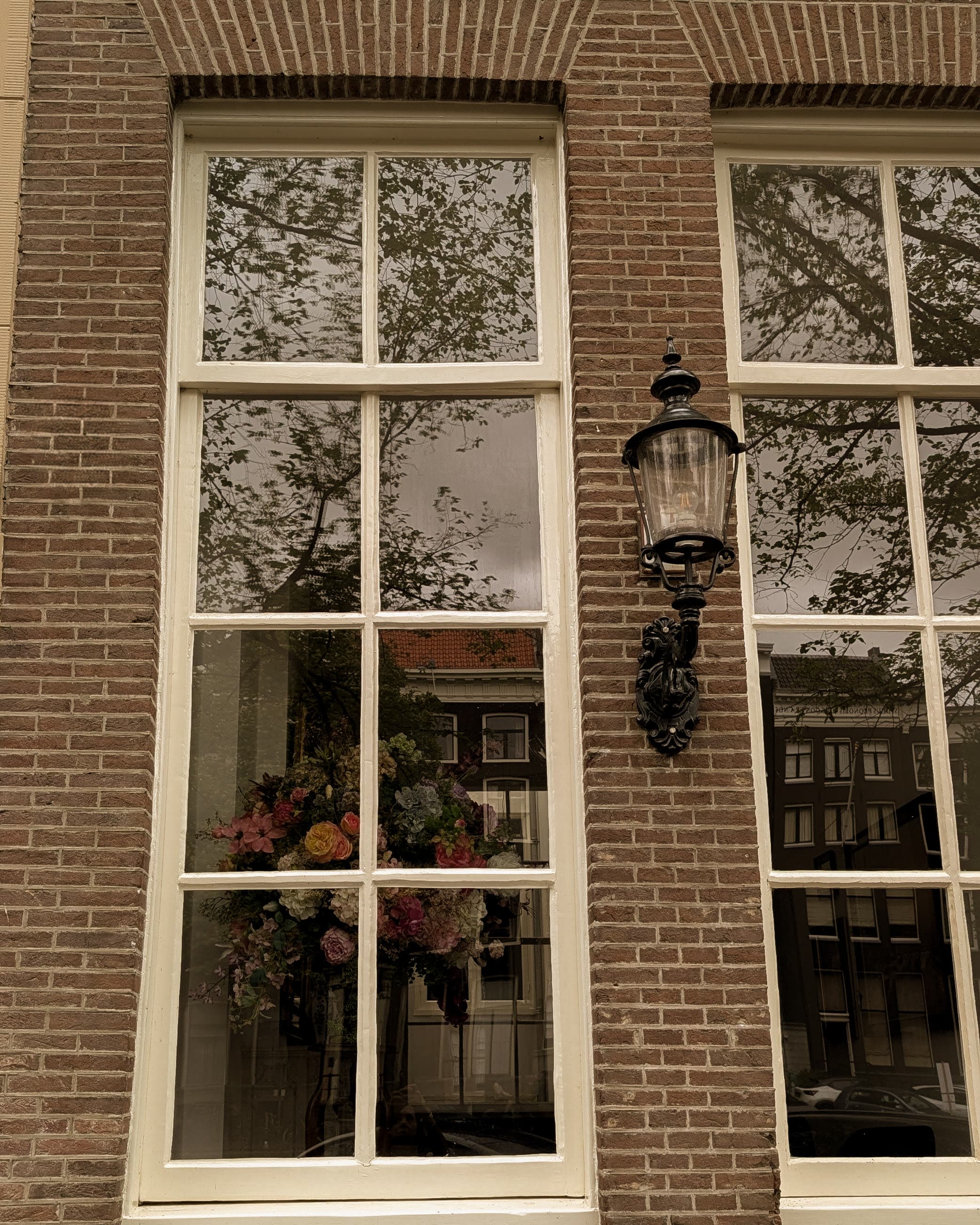 A lit Amsterdam canal house window at dusk with a floral arrangement on the sill
