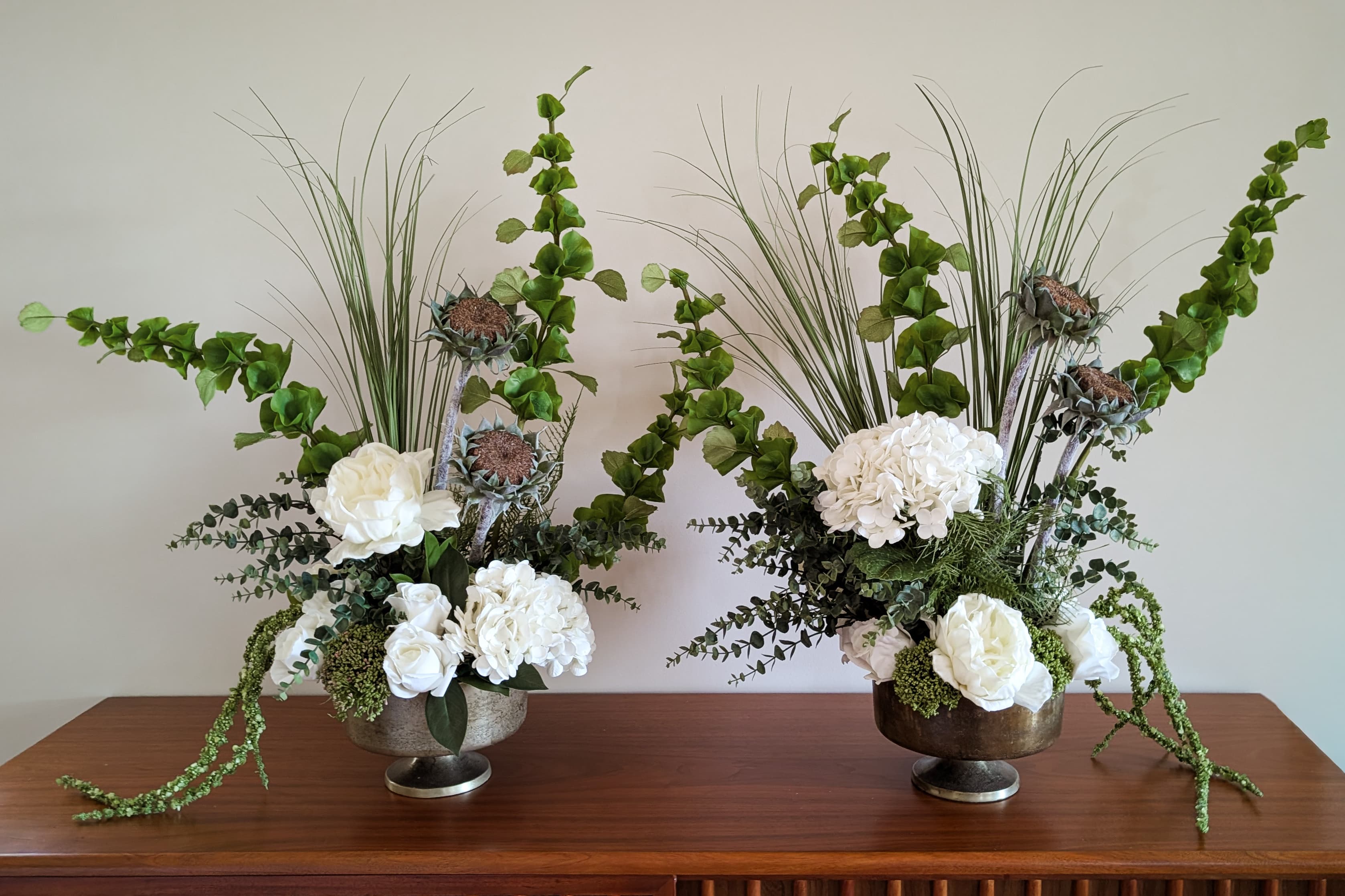 Two Atelier Bloem forever floral arrangements in bronze pedestal vessels, with white hydrangea, garden roses, eucalyptus, ginkgo vine, thistle, and ornamental grasses
